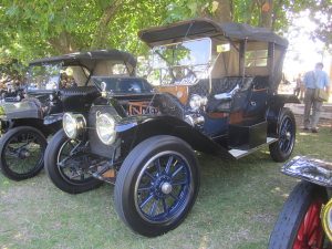 Vuoden 1912 Cadillac Tourer on tummansininen kanttiauto, jossa on pinnapyörät ja ylös nostettu kuomu.