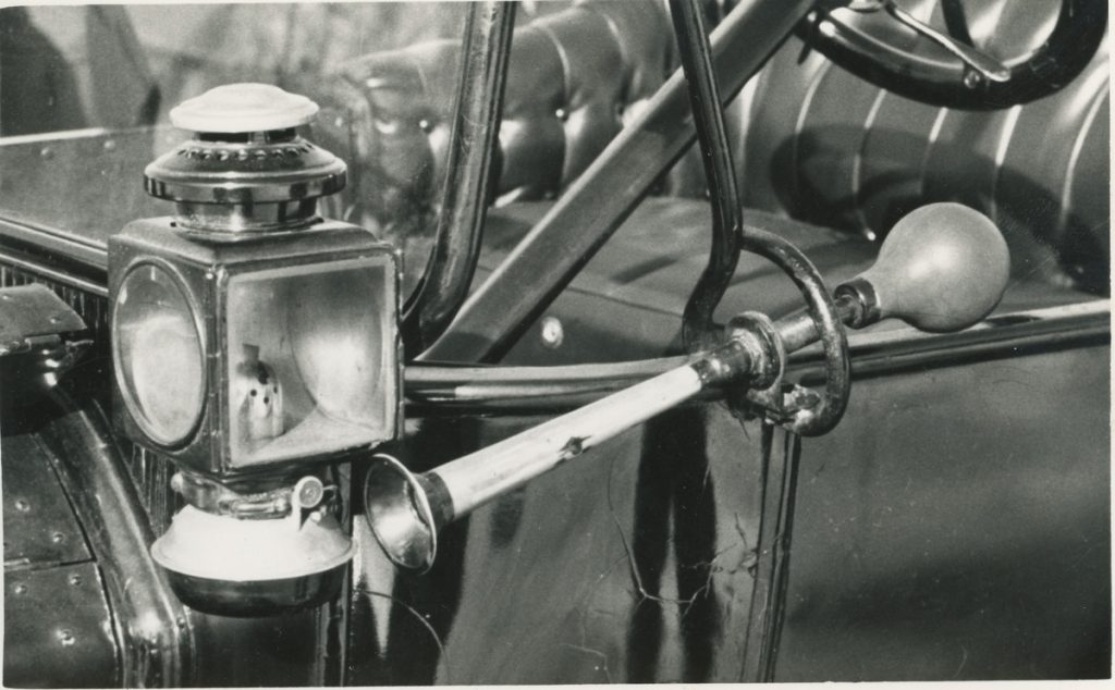 Mustavalkoisessa kuvassa on etualalla kuutionmallinen autonlyhty, sen alla pitkävartinen töötti-torvi ja taustalla näkyy avointa 1900-luvun alkukymmenten henkilöauton koria.