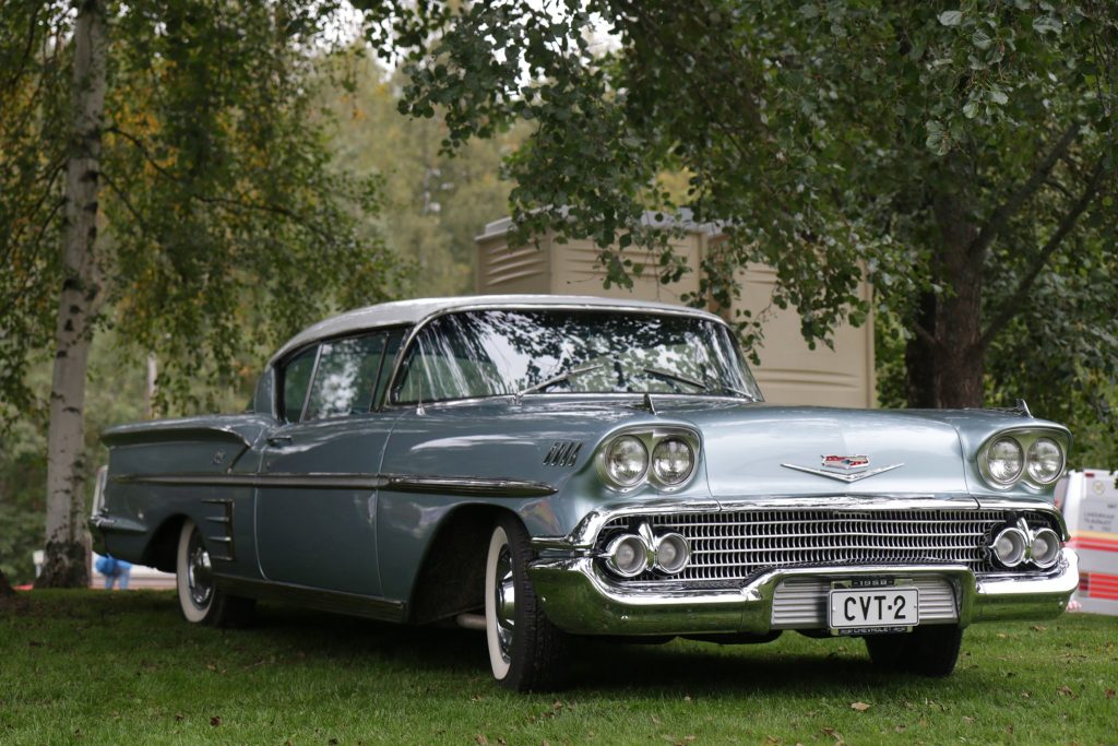 Metallinhohtoinen Chevrolet Impala vuodelta 1958 nurmikolla puiden siimeksessä. 
