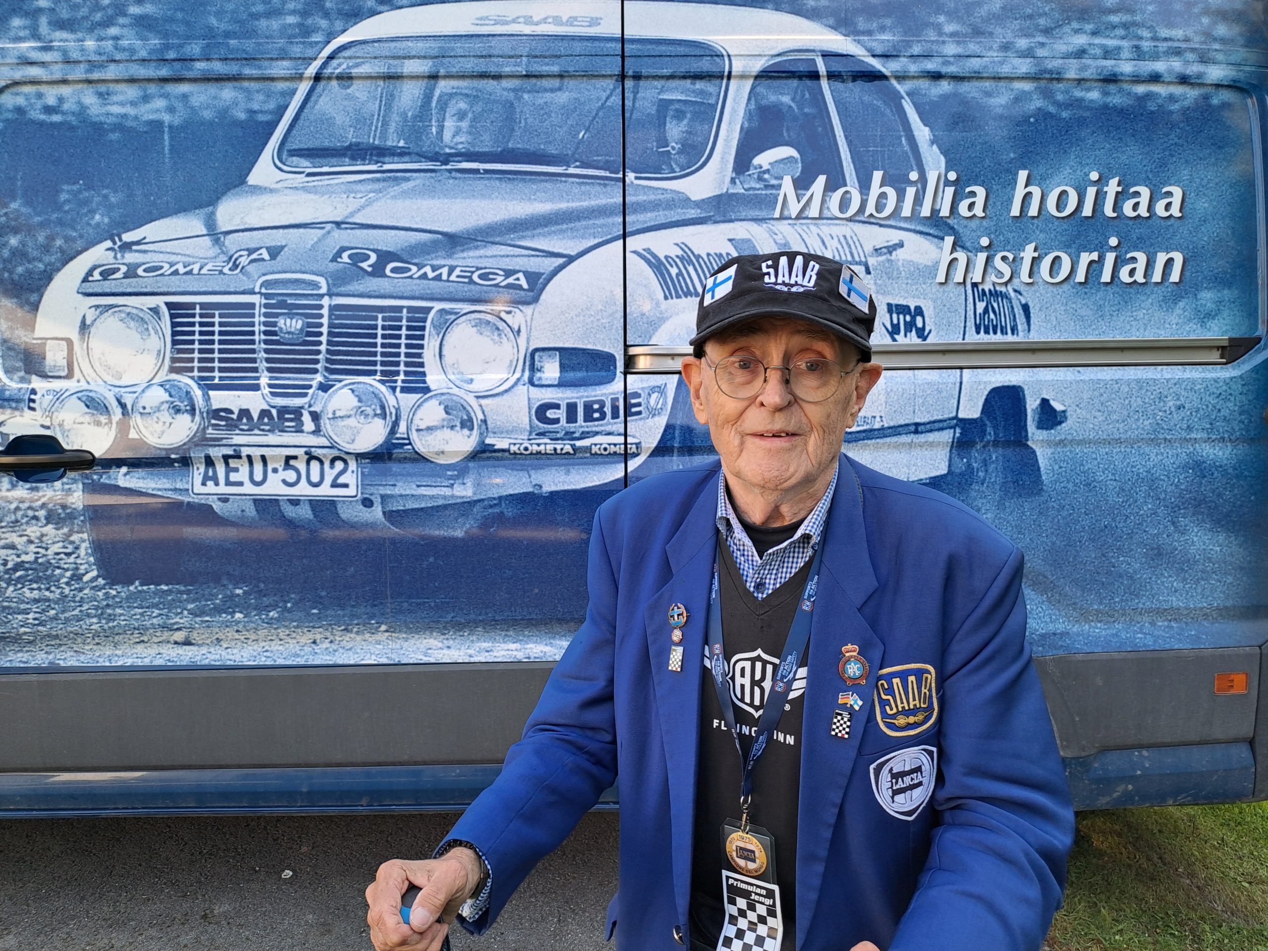 80-vuotias Simo Lampinen sininen takki ja lippalakki päässään sinivalkoisen rallikuvan edessä.