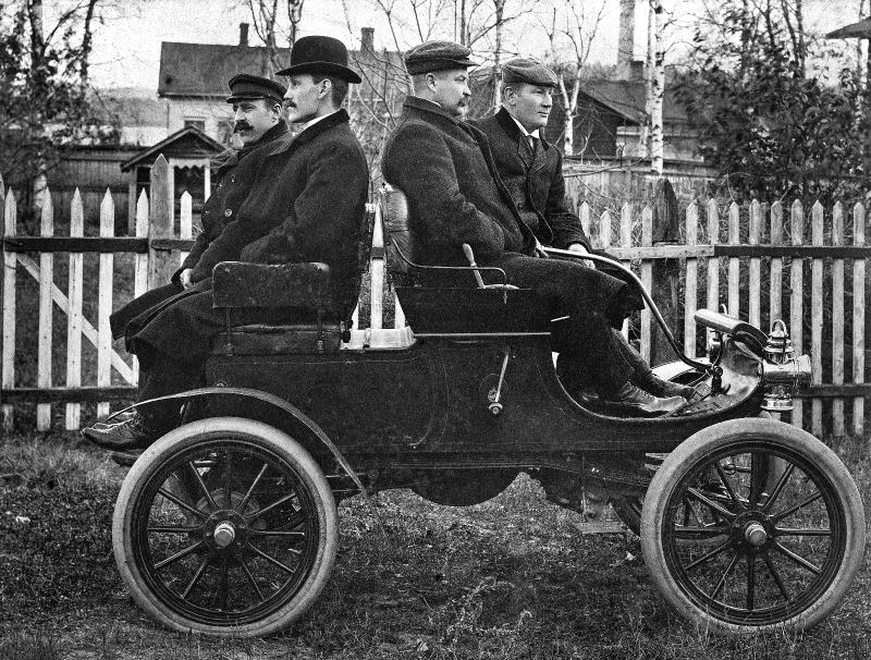 Mustavalkoisessa kuvassa 1900-luvun alusta hevosvaunun näköisen auton kyydissä istuu miehiä. Kuljettaja ja repsikka katsovat eteenpäin, matkustajat istuvat selkä menosuuntaan päin takapenkillä.
