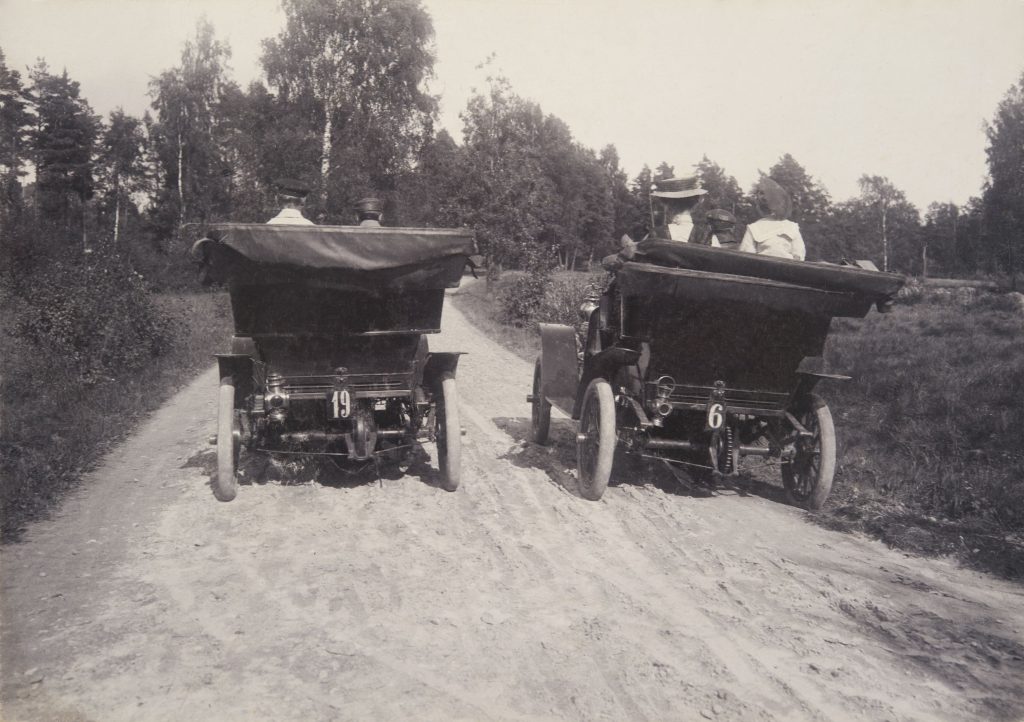 Mustavalkokuvassa kaksi 1910-luvun autoa vierekkäin hiekkatiellä kuomut alaslaskettuina ajamassa poispäin kuvaajasta. Autojen takaosassa on numerokyltit 19 ja 6, jotka ovat varhaiset rekisterikilvet.