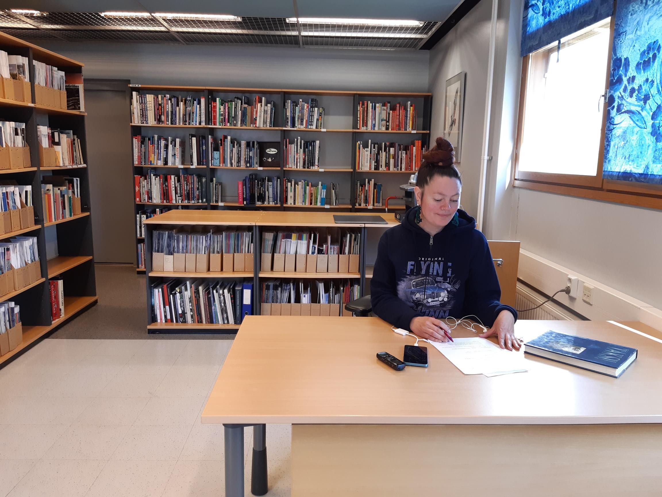 Nainen korkea nuttura päälaella istuu pöydän ääressä tekemässä kirjallista työtä, takana näkyy kirjaston kirjahyllyjä.