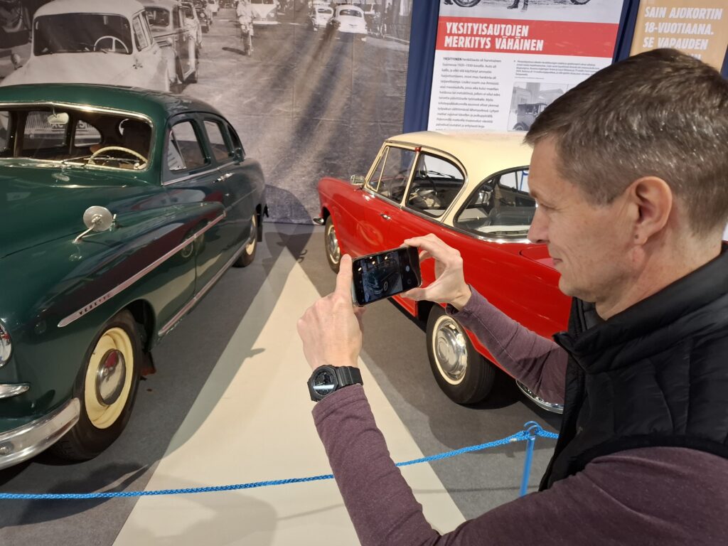 Mies kuvattuna olan takaa ottamassa valokuvaa kännykällä vihreästä museoautosta automuseossa.