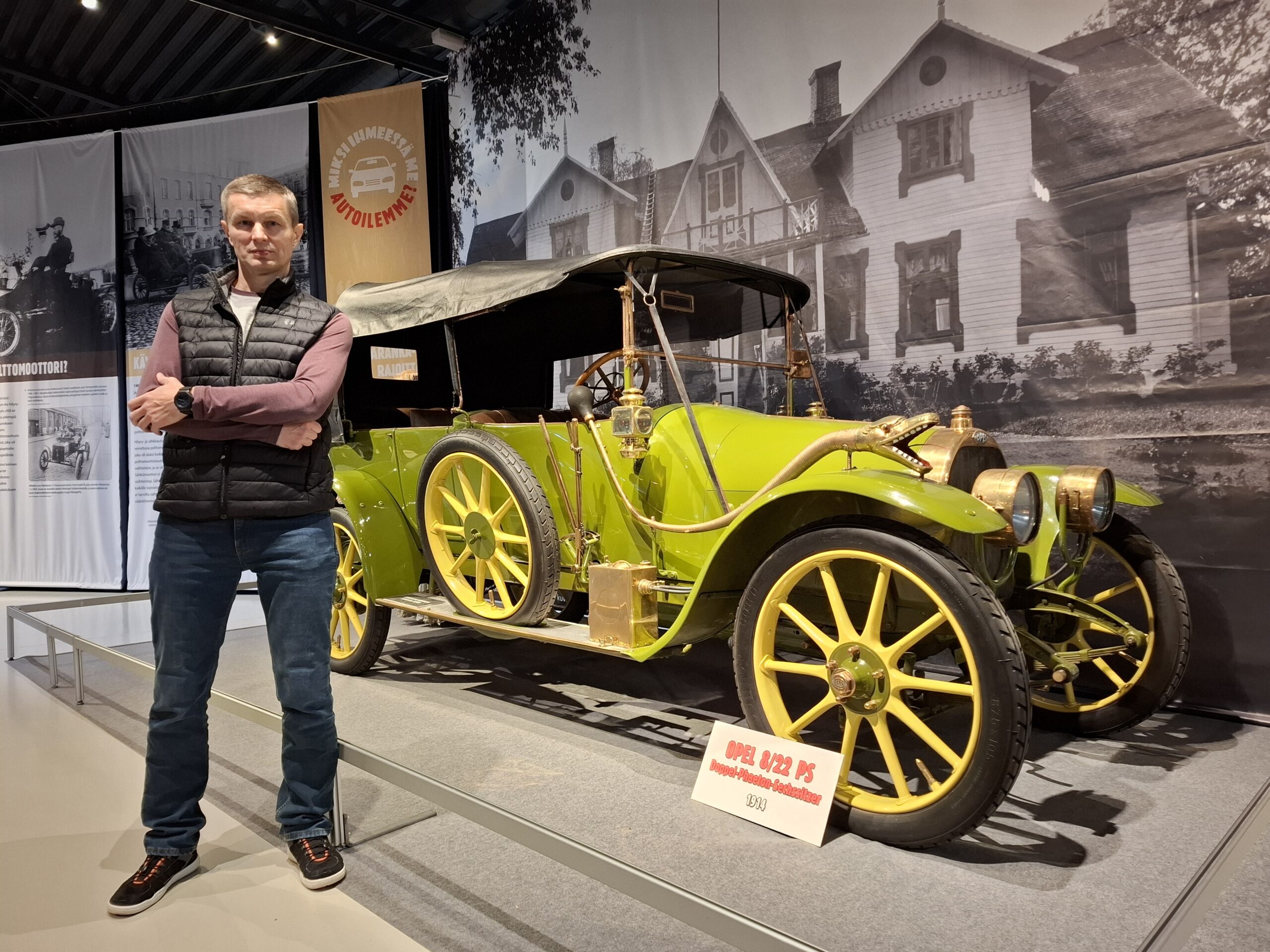 Mies seisoo kädet puuskassa 1900-luvun alun henkilöauton vieressä automuseossa.