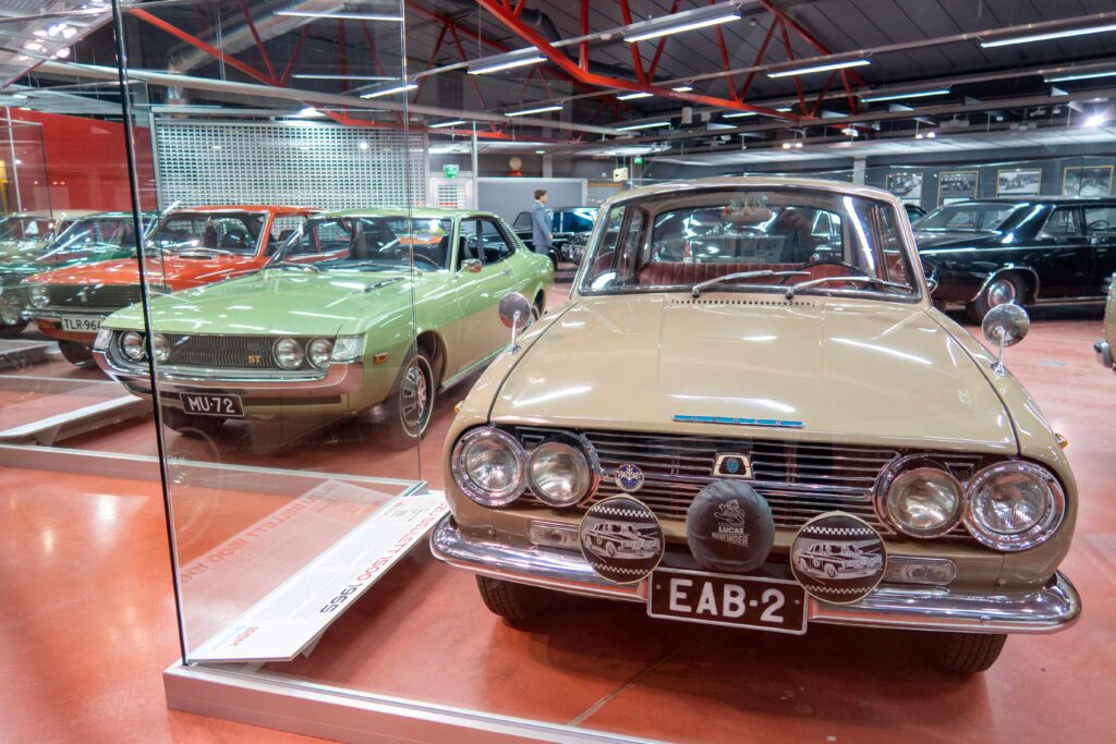 Vaaleanbeige Isuzu Bellet lisäajovaloineen kuvattuna edestäpäin näyttelytilassa. Taustalla näkyy muita näyttelytilan ajoneuvoja.