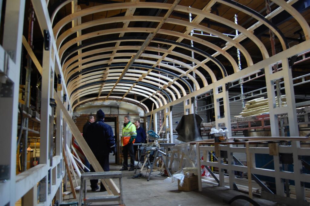 Kaarevan kehikon alla rakennustarvikkeita ja taka-alalla neljä ihmistä.