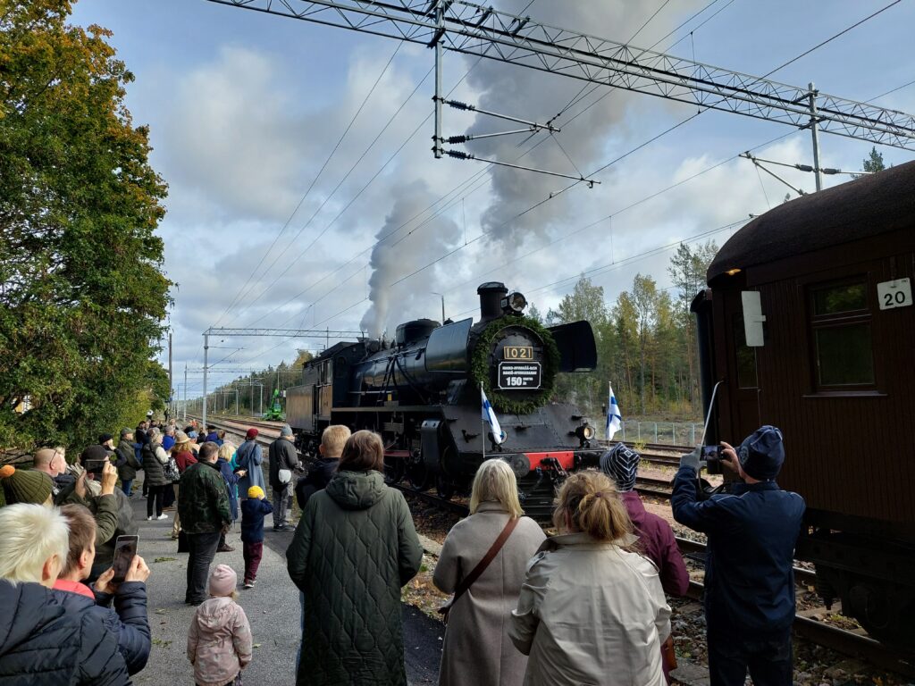 Höyryveturi koristeltuna Suomenlipuin, etualalla katselijoita.