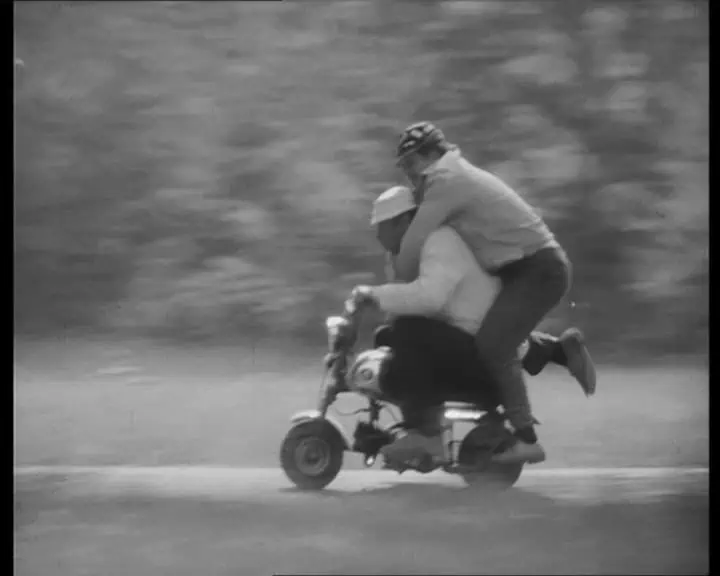 Mustavalkoinen kuva, jossa kaksi aikuista huristelee pienen pienellä mopolla kesämaisemassa. Toinen istuu mopon satulassa ja toinen yrittää pysyä kyydissä seisten ja kaverin kaulasta kiinni pitäen.