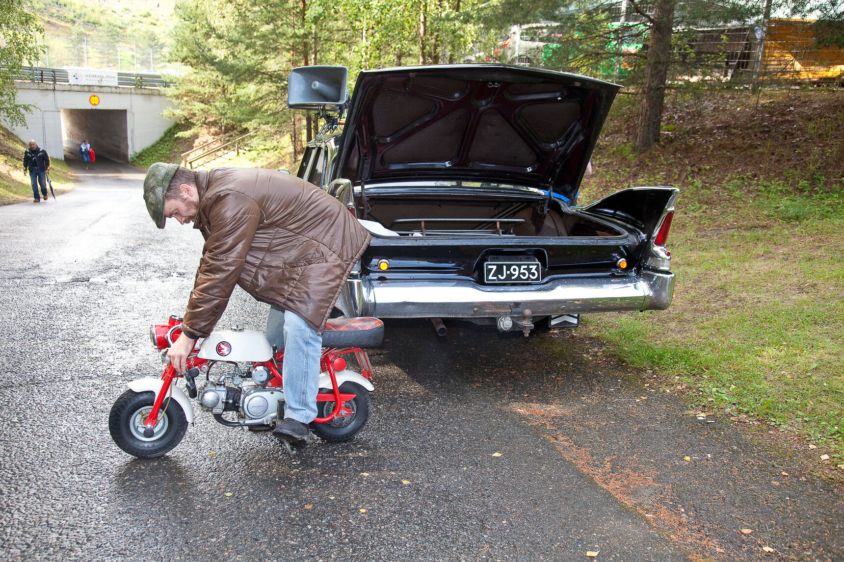 Mies pienen punavalkoisen Monkey-mopon päällä, taustalla musta Plymouth iso takaluukku avoinna.