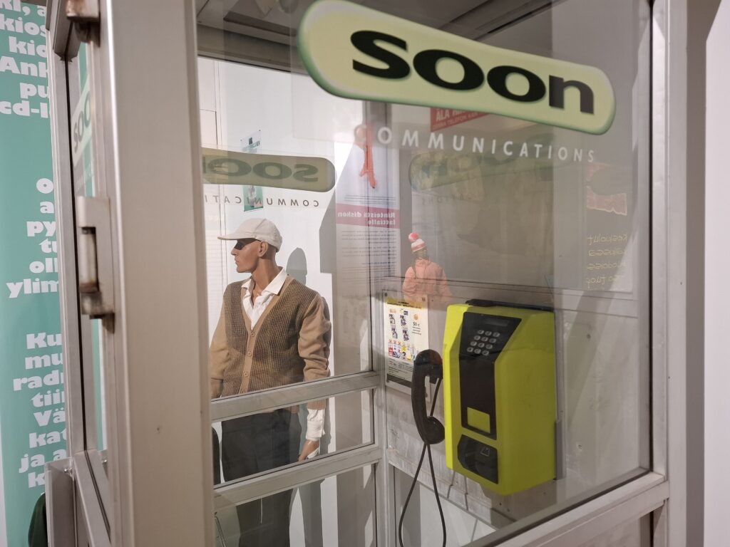 Kuva on otettu Soon-teippauksissa olevan puhelinkioskin läpi. Kioskissa näkyy musta-keltainen puhelin ja kopin takana lippalakkipäinen mallinukke.