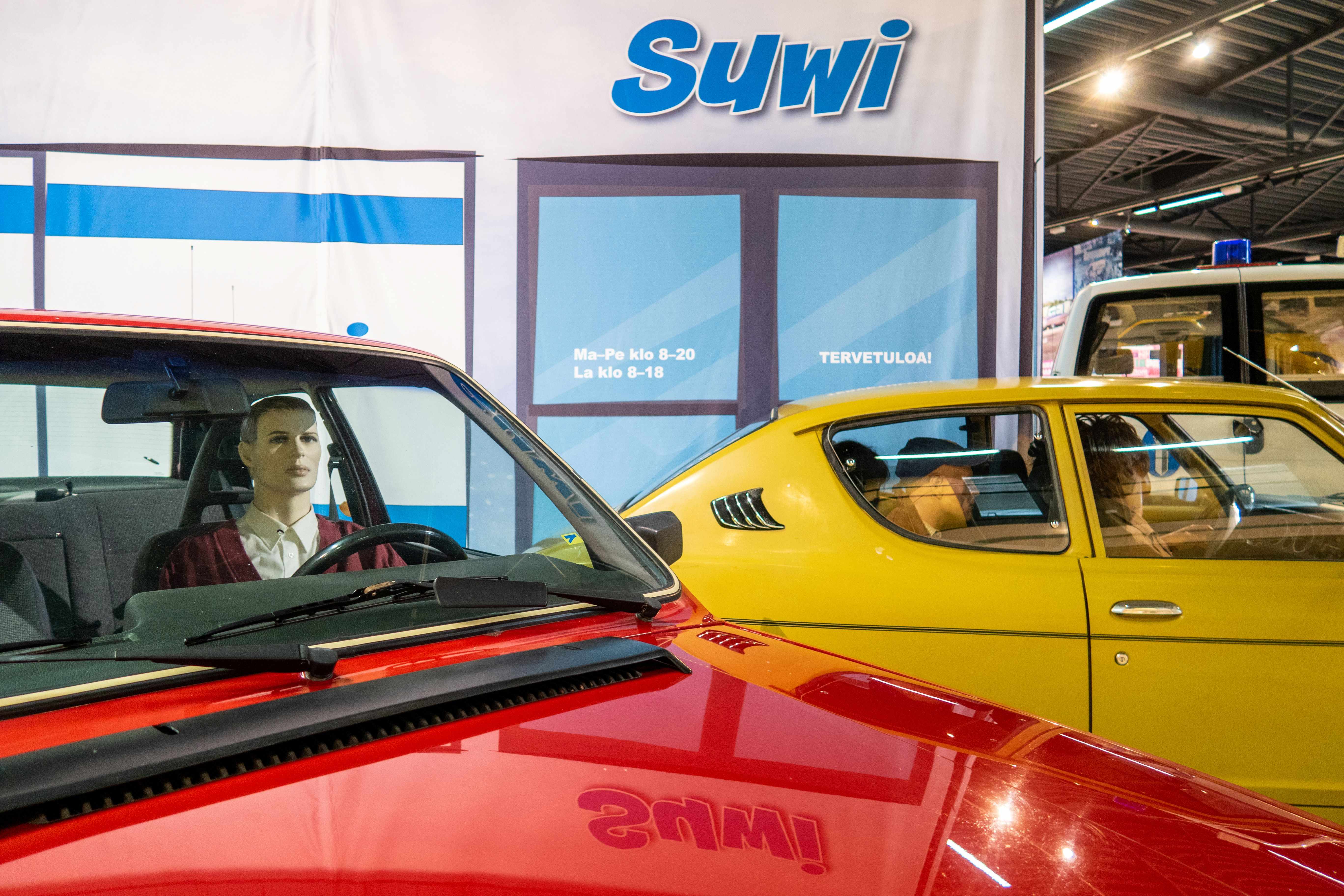 Näyttelyseinäkkeen edessä punainen Volvo kuskin kanssa ja taustalla kellertävän ruskea Datsun 100 A täydessä matkustajalastissa.