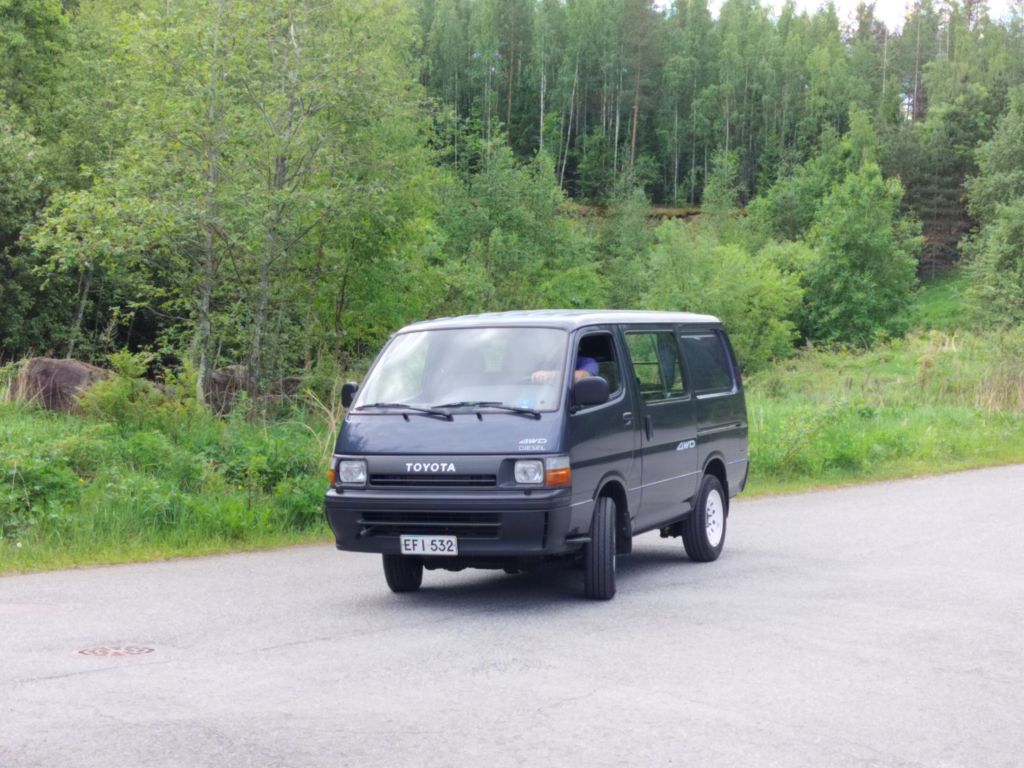 Mustansinertävä Toyota Hiace kuvattuna etuviistosta asfaltilla metsätaustanaan.